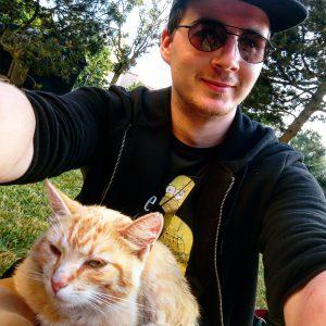 Felix T. Vogel sitzt mit einer Sonnenbrille und Katze auf einer Wiese