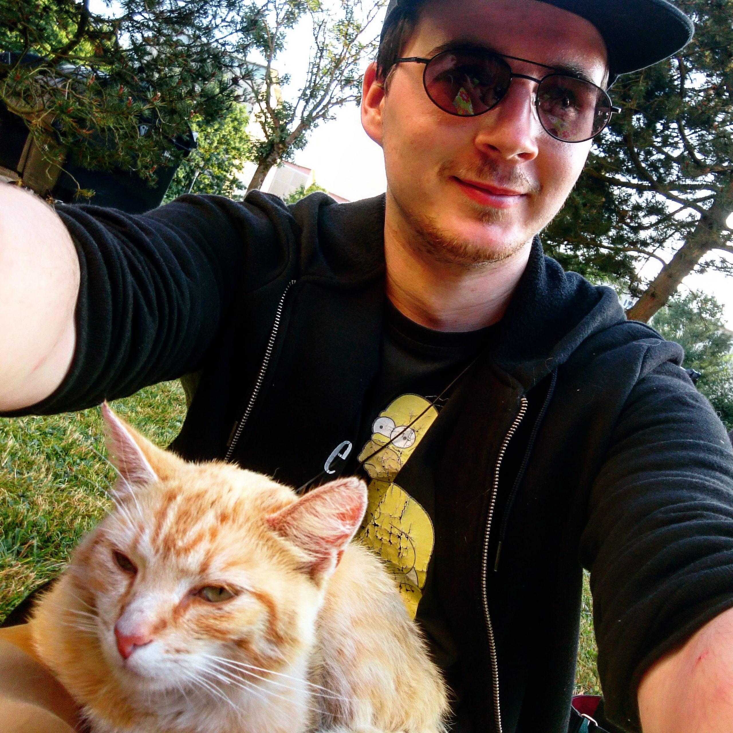 Felix T. Vogel sitzt mit einer Sonnenbrille und Katze auf einer Wiese